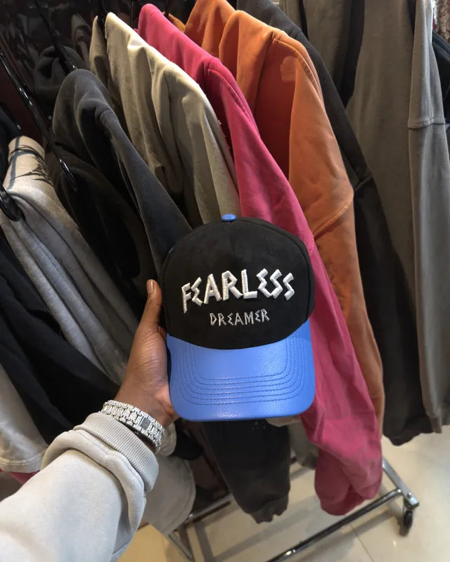A Fearless Dreamer cap held up in front of a clothing rack